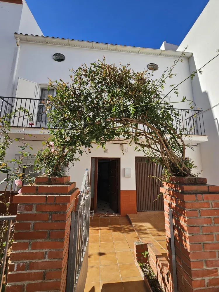 Edificio blanco de dos pisos con fachada de ladrillo y balcón, entrada enmarcada por plantas trepadoras, día soleado.