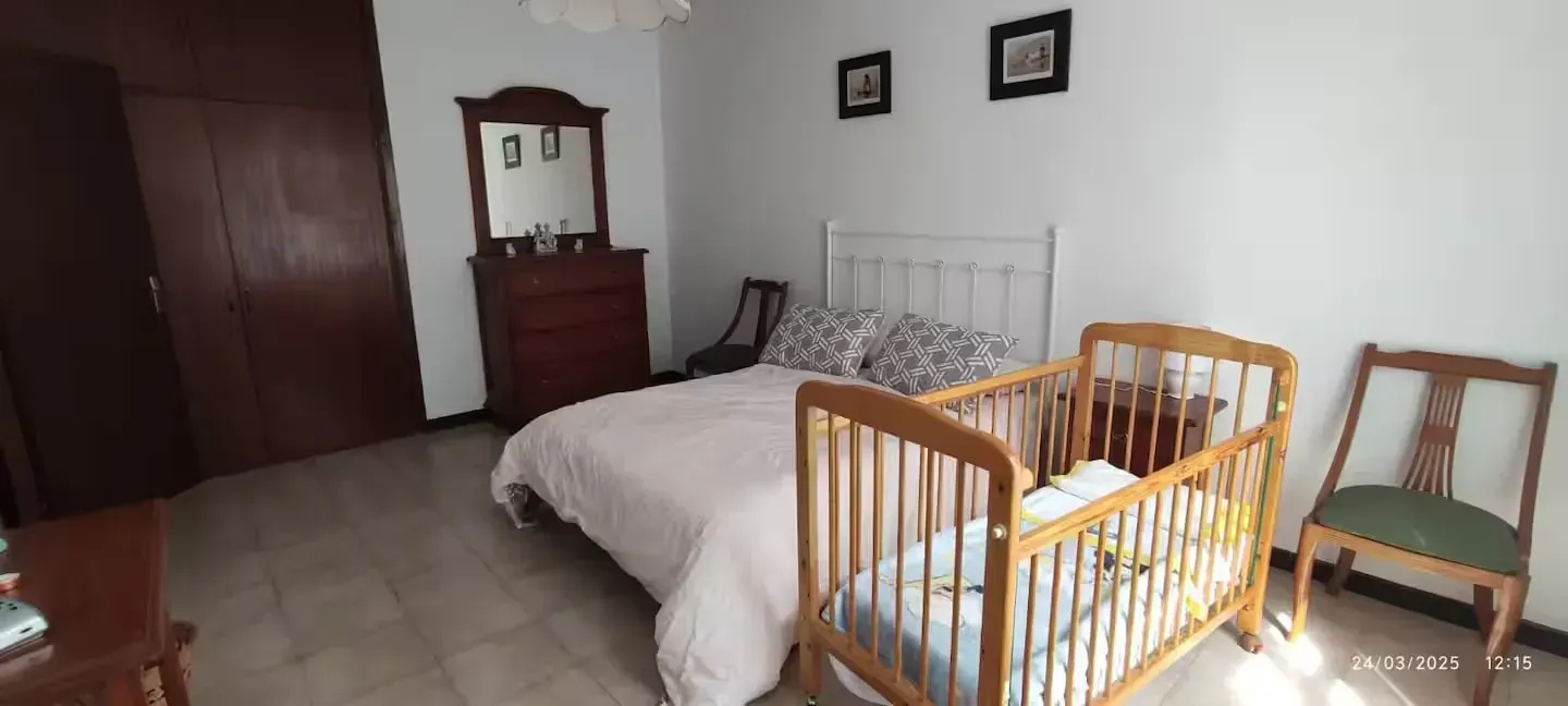 Dormitorio con cuna de madera, cama, cómoda con espejo y dos pequeños cuadros enmarcados.