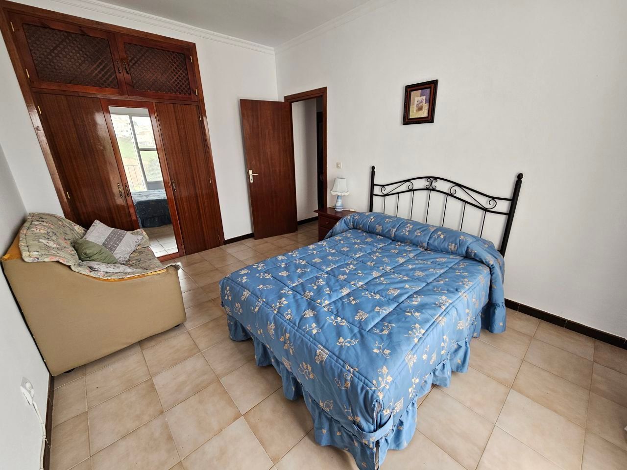 Dormitorio con cubrecama floral azul, marco de cama de hierro forjado, piso de baldosas color canela y una puerta abierta.