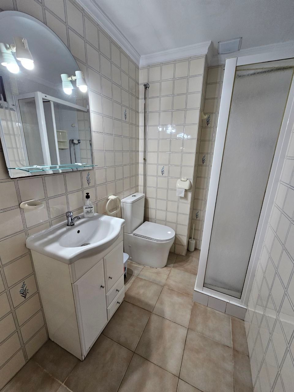 Baño con lavabo, inodoro y ducha blancos. Paredes y suelo de azulejos beige.