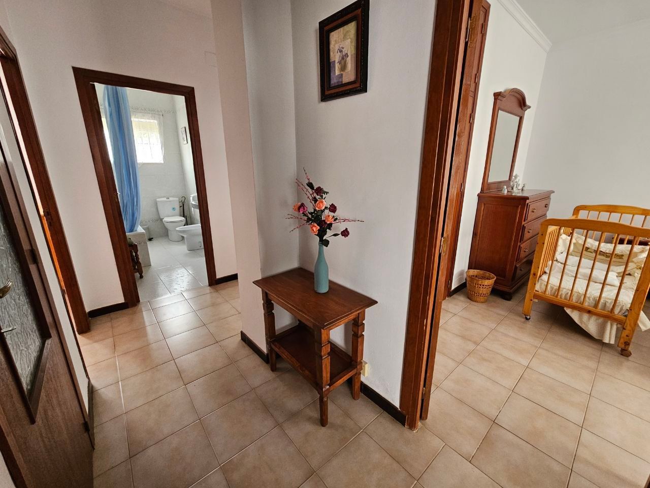 Pasillo con puertas que dan a un baño y un dormitorio. Mesa de madera con jarrón, obras de arte y muebles.