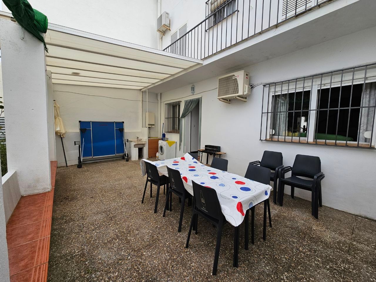 Patio al aire libre con mesa y sillas bajo un dosel, contenedor de almacenamiento azul y aire acondicionado en la pared.
