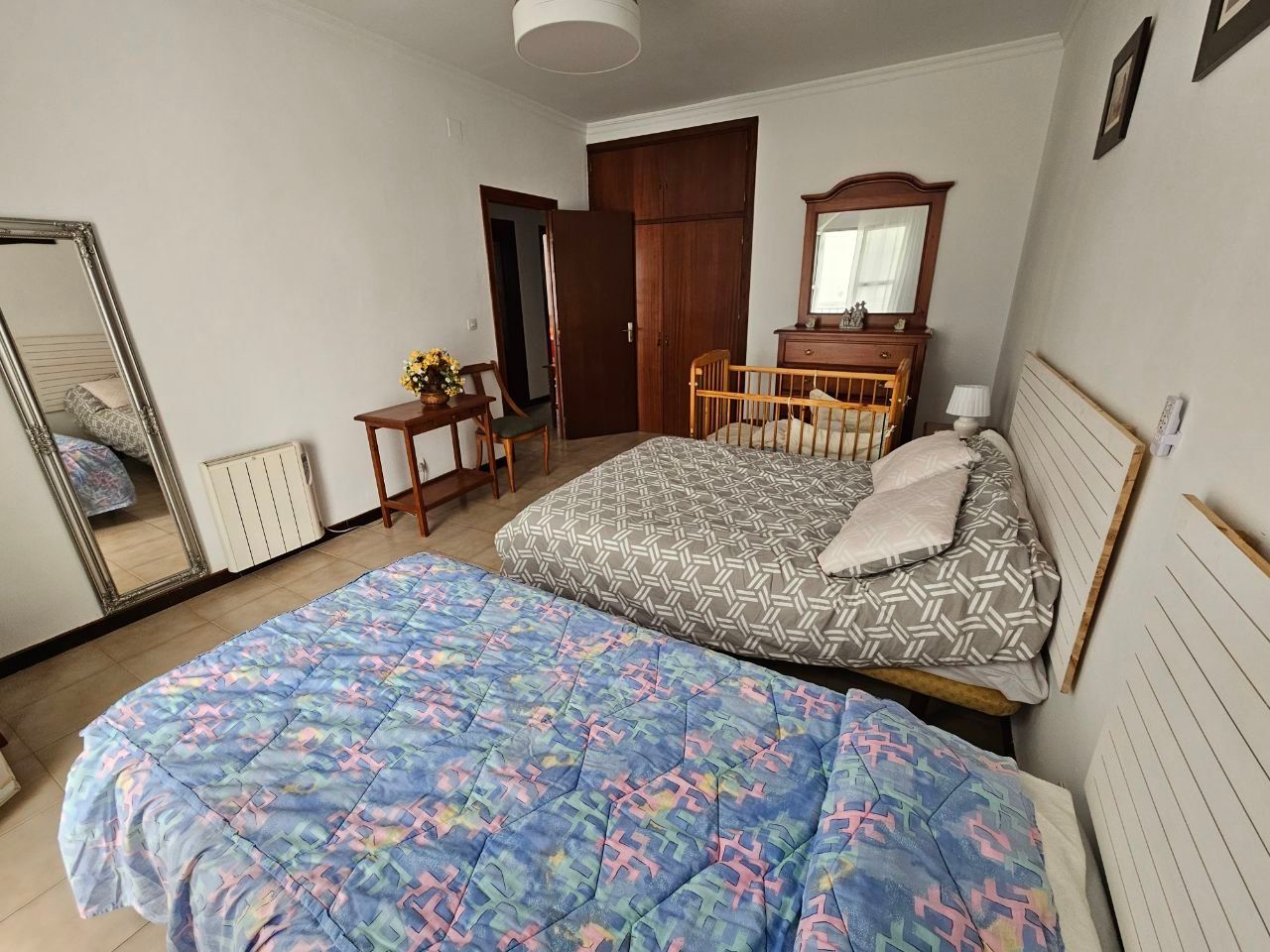 Dormitorio con dos camas, cuna, cómoda, espejo y armario; paredes blancas, tonos neutros.