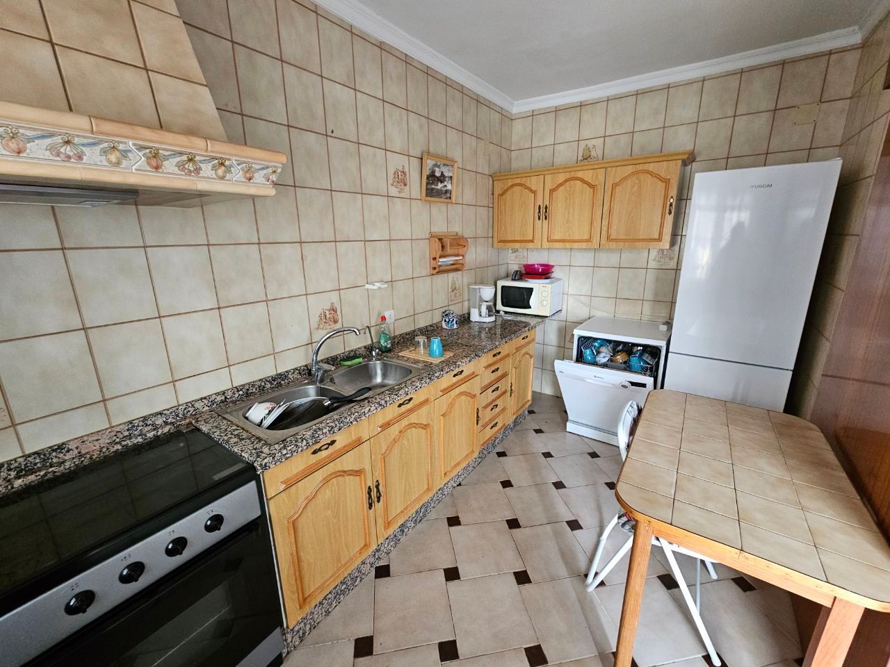 Cocina con gabinetes, estufa, fregadero, refrigerador, microondas y mesa. Paredes de azulejos beige y piso a cuadros negros y beige.