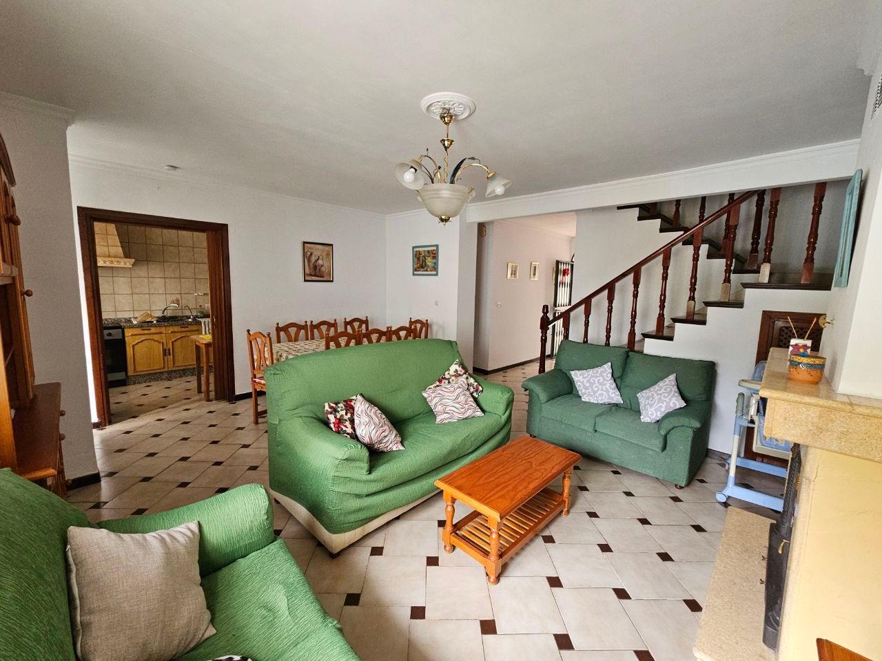 Sala de estar con sofás verdes, mesa de centro de madera y escalera. Cocina visible a través de la puerta.