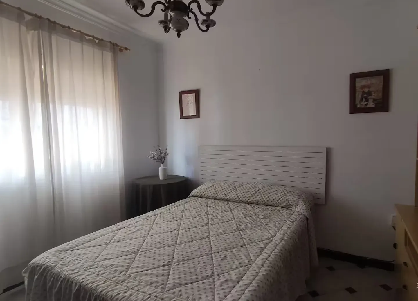Dormitorio con una cama individual, una pequeña mesa redonda, arte enmarcado, cortinas transparentes y una lámpara de araña.