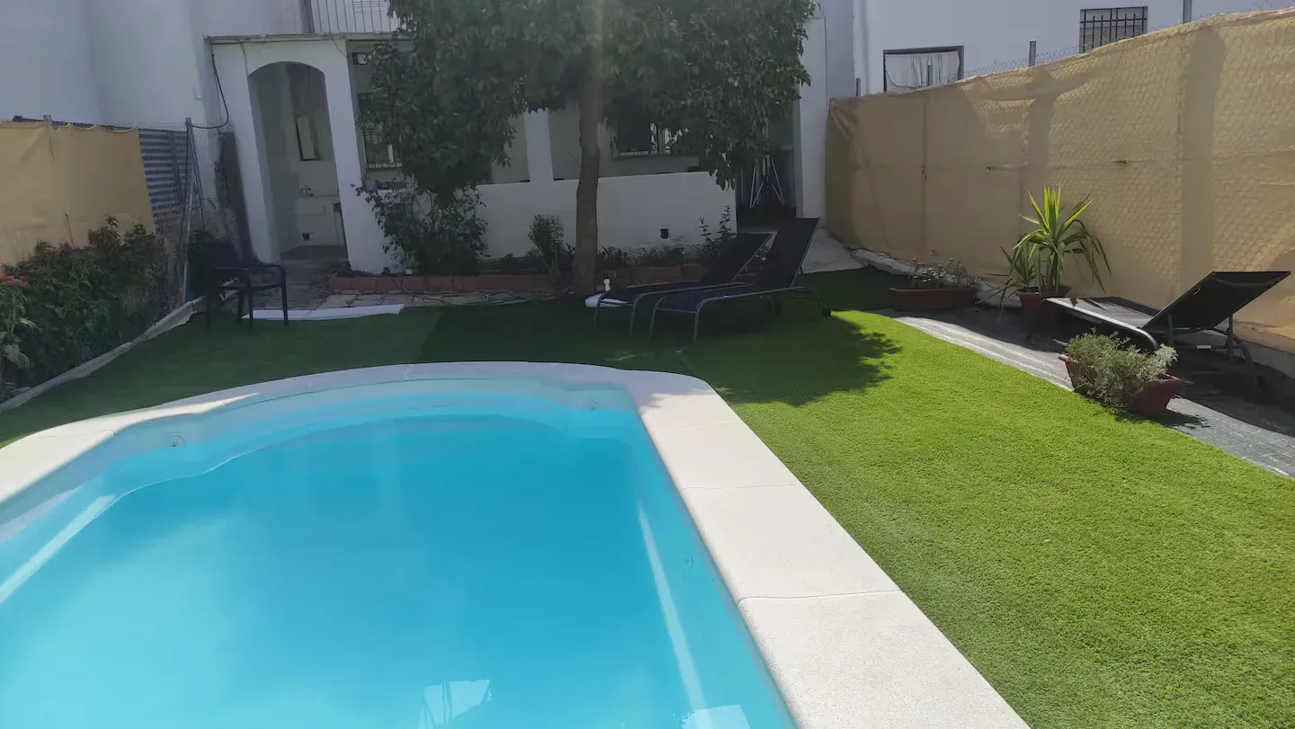 Vista a la piscina, con césped, árboles y tumbonas. Día soleado y luminoso.