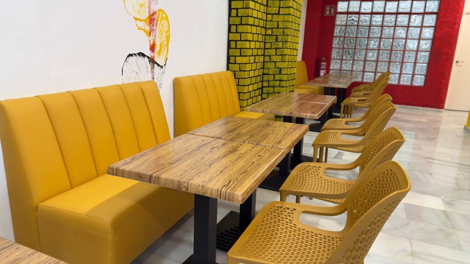 Asientos amarillos en cabinas y sillas de mimbre junto a mesas de madera en el luminoso interior de una cafetería o restaurante.