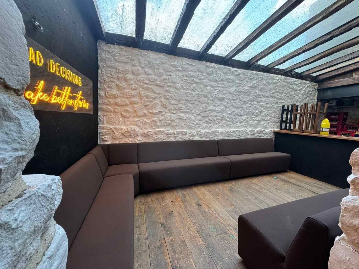 Salón moderno al aire libre con sofás modulares oscuros, paredes de piedra y techo de cristal cubierto con un letrero de neón.