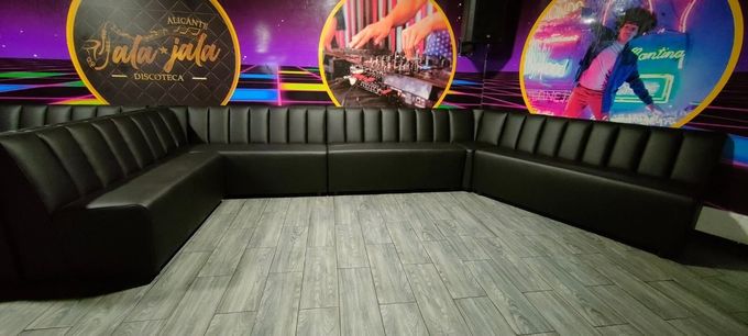 Sala de estar moderna con asientos negros en forma de L, suelo de madera y murales coloridos al fondo