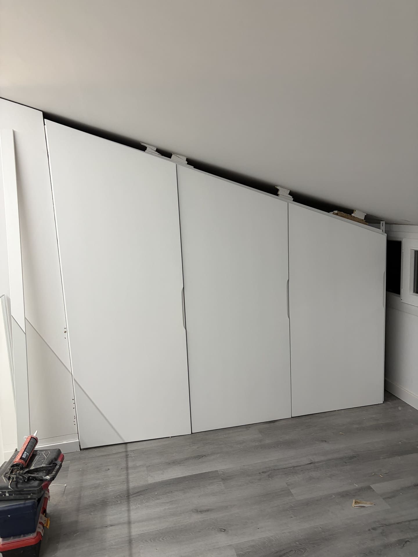 Armarios empotrados blancos instalados bajo un techo inclinado, junto a suelo gris. Se ven herramientas de construcción.