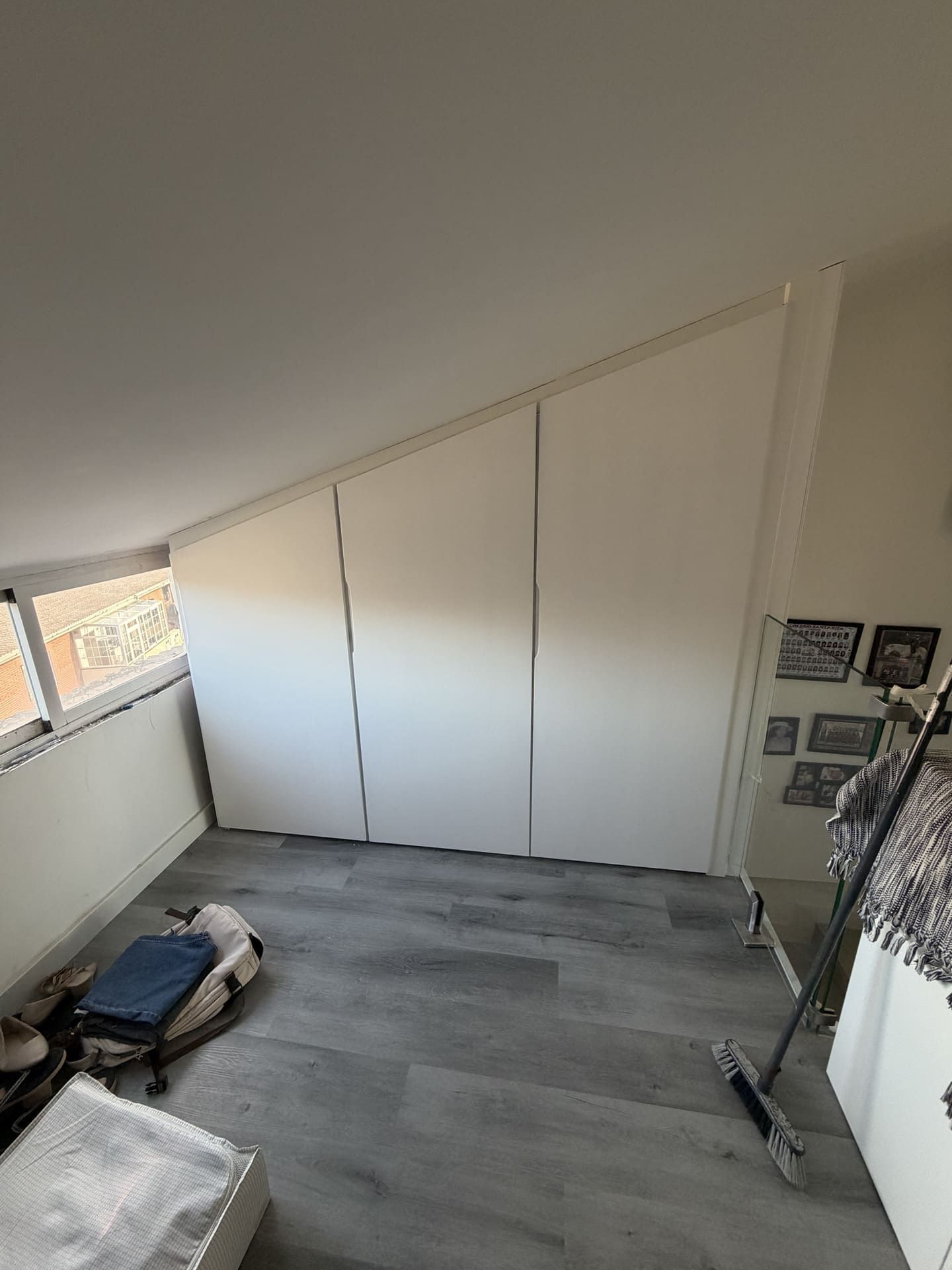 Puertas de armario blancas inclinadas en una habitación abuhardillada con suelo gris y una ventana.