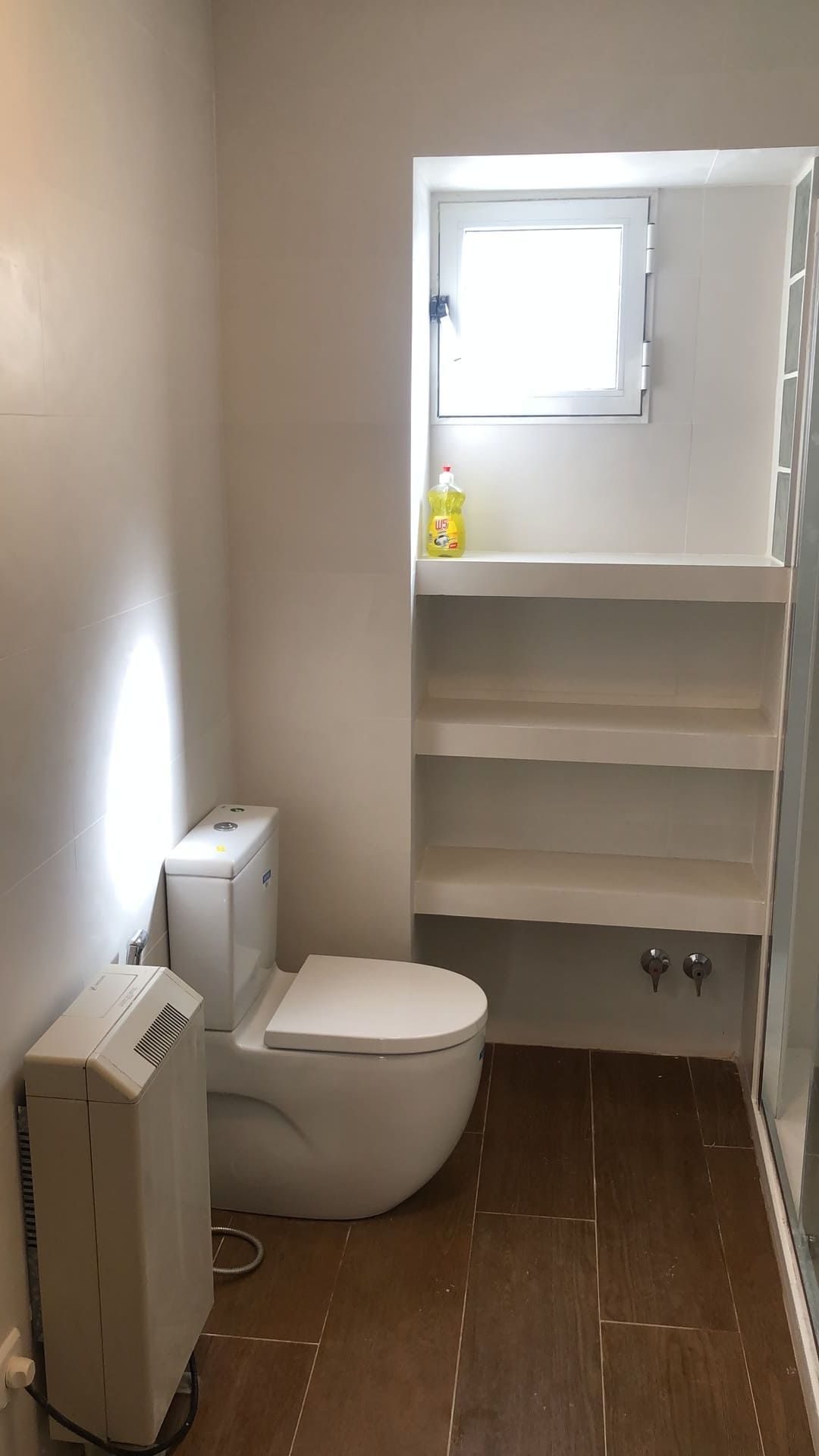 Baño pequeño con inodoro, estanterías y una pequeña ventana. Suelo de baldosas marrones y paredes blancas.