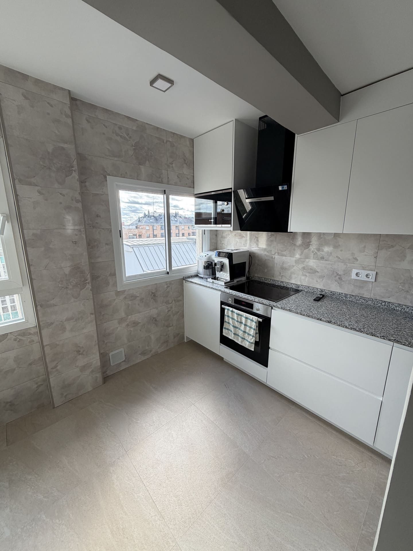 Cocina moderna con gabinetes blancos, encimeras grises y paredes de azulejos con efecto piedra. Electrodomésticos negros y una ventana.