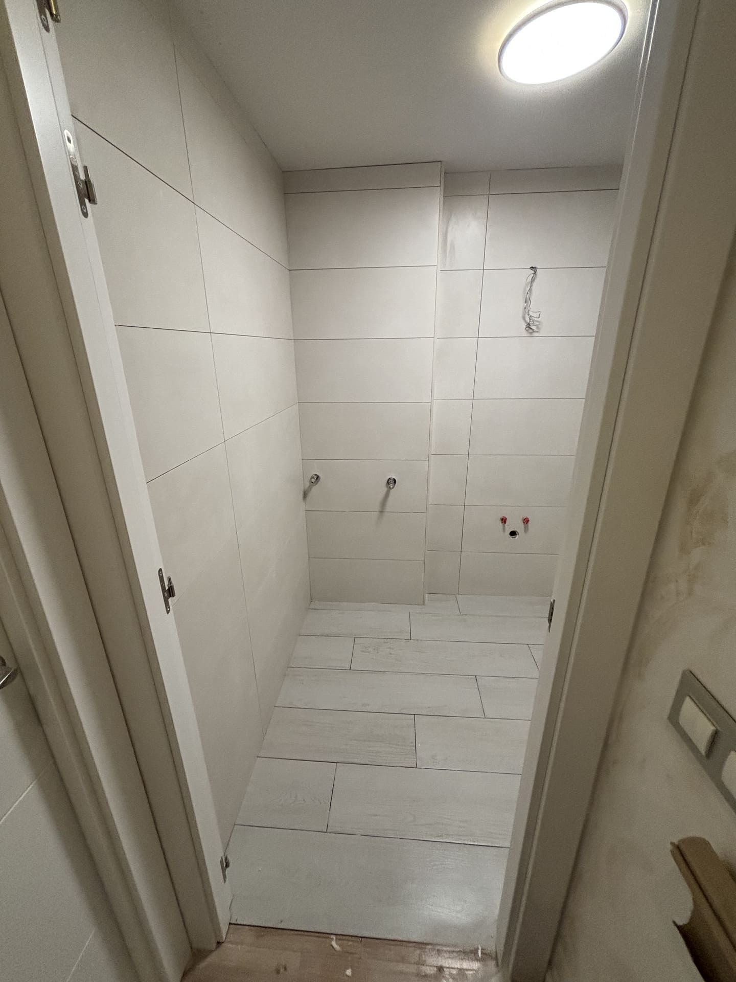 Pequeño baño en construcción; paredes y piso de azulejos blancos, accesorios instalados, luz de techo.