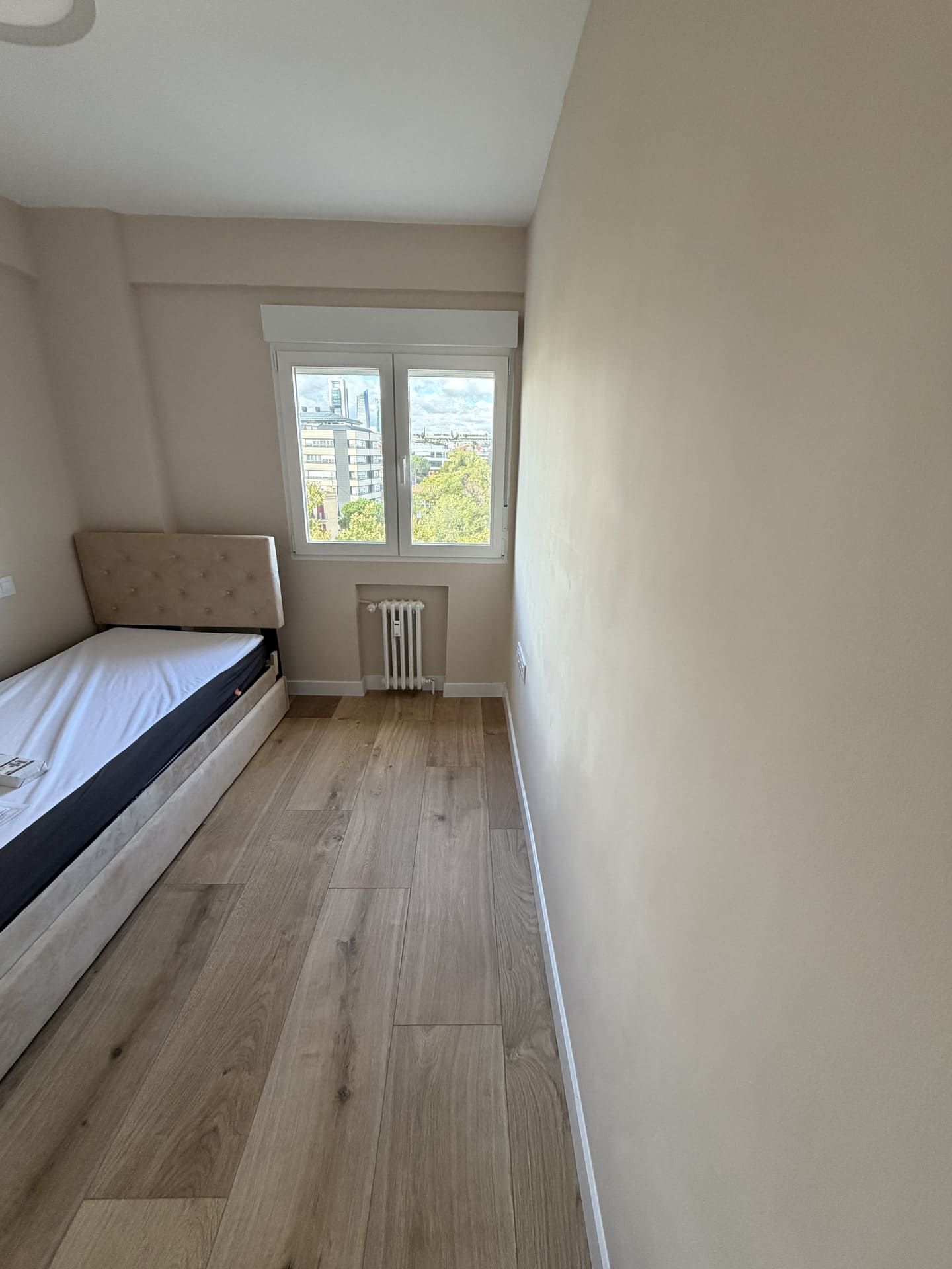 Pequeño dormitorio con una cama individual junto a una ventana, pisos de madera clara y paredes beige.