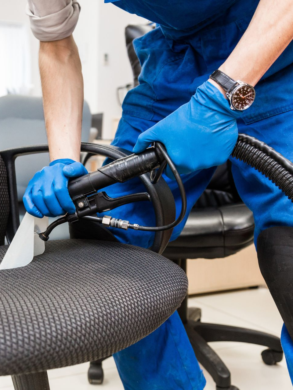 Une personne en blouse bleue nettoie une chaise de bureau noire avec un outil de nettoyage, portant des gants bleus.