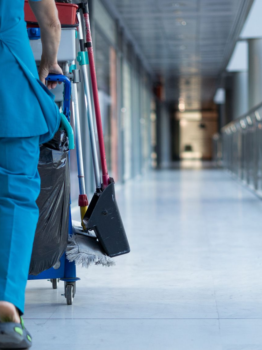 Une personne en blouse bleue pousse un chariot de nettoyage dans un couloir d'hôpital.
