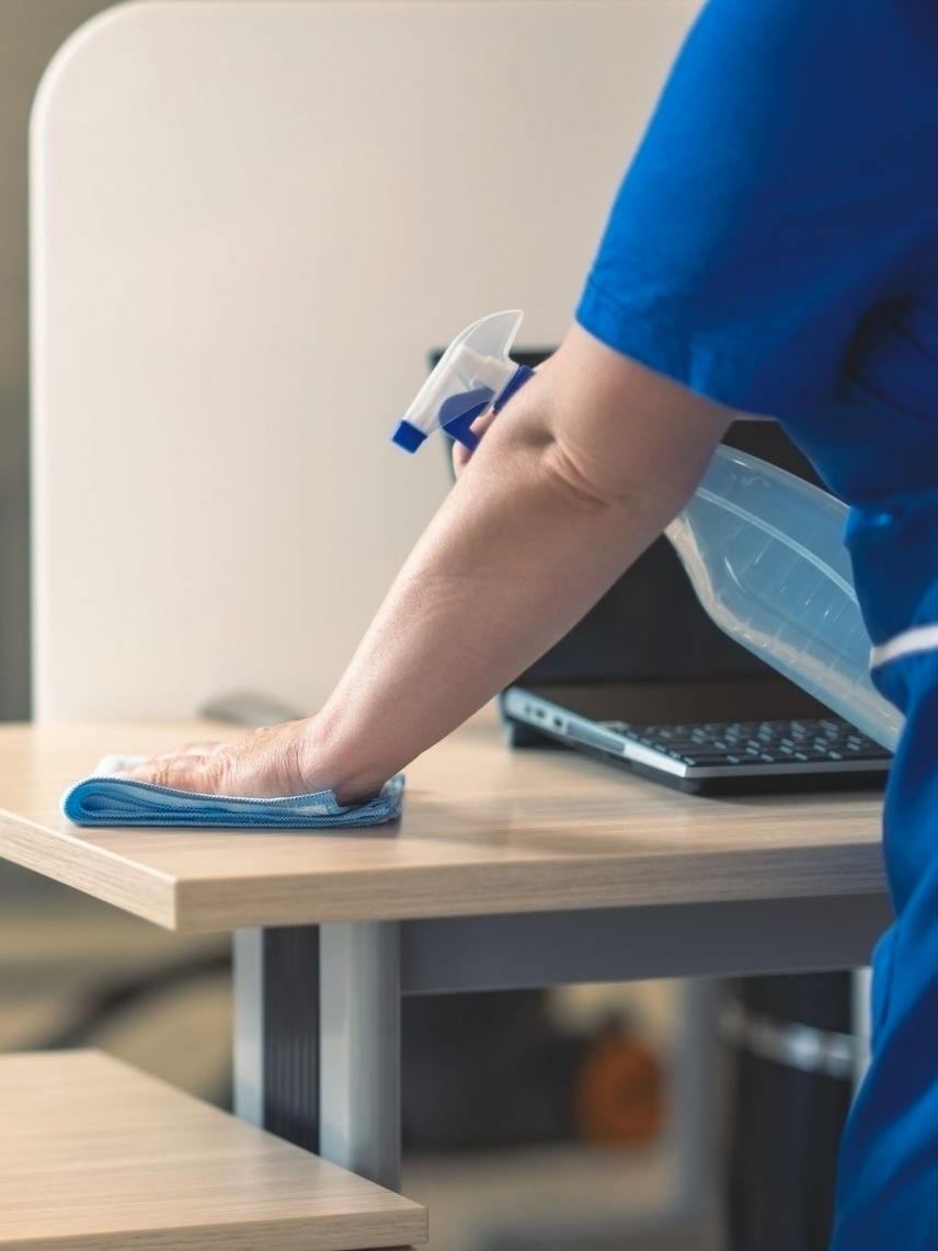 Une personne en blouse bleue désinfecte un bureau en bois clair avec un vaporisateur et un chiffon.