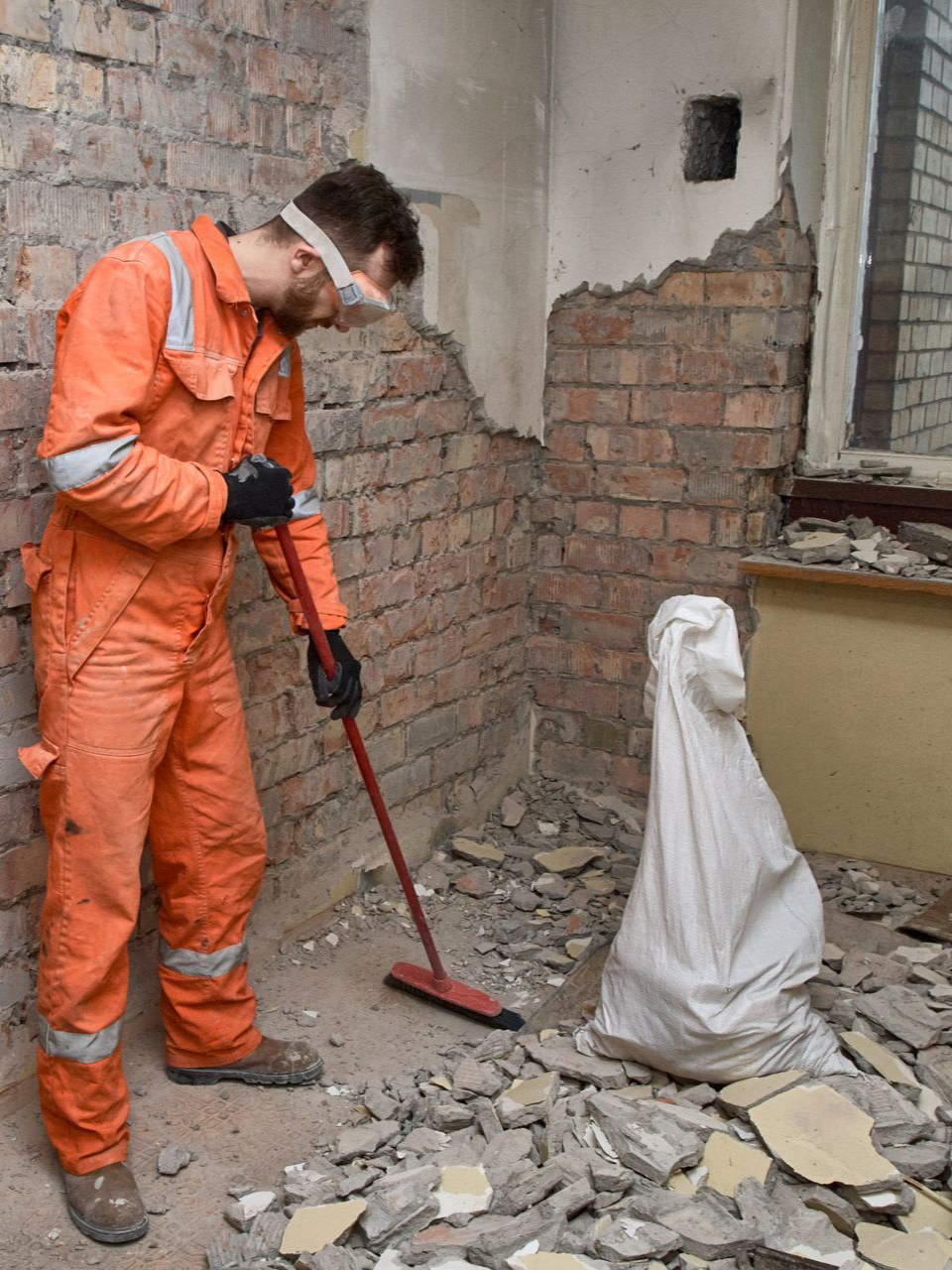 Une personne en combinaison orange balaie des débris dans une pièce aux murs de briques apparentes.