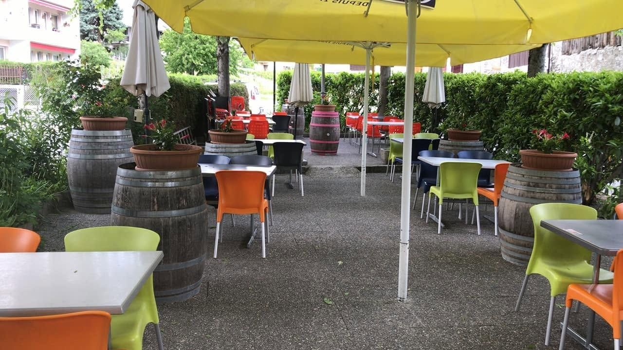 Terrasse de café en plein air avec parasols jaunes, chaises colorées et tonneaux en bois entourés de verdure.