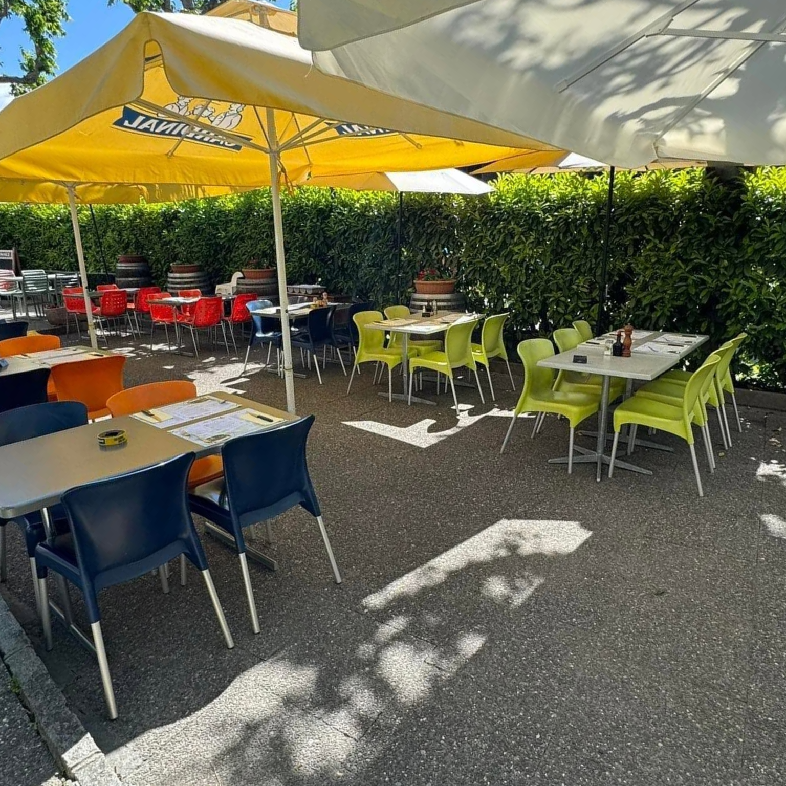 Terrasse ensoleillée avec terrasse de café ombragée par des parasols jaunes et blancs, à côté d'une haie.