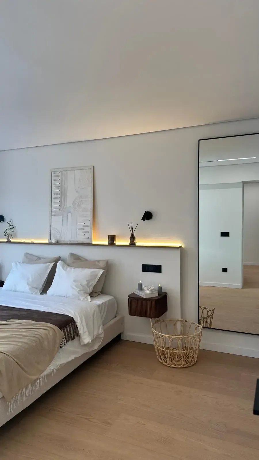Dormitorio minimalista con ropa de cama blanca, suelo de madera, cuadros en la pared, lámpara de noche, espejo y cesta