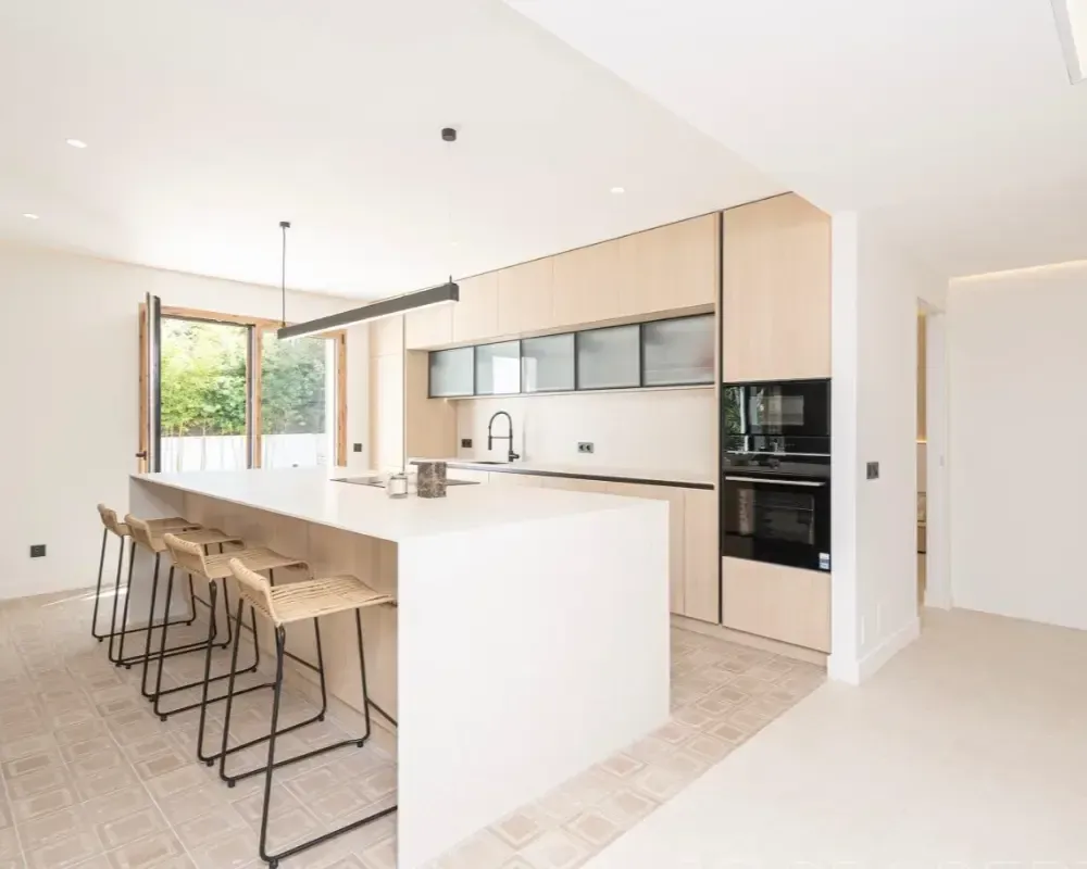 Cocina moderna y luminosa con isla blanca, armarios de madera, taburetes de bar y mesa de comedor junto a la ventana.