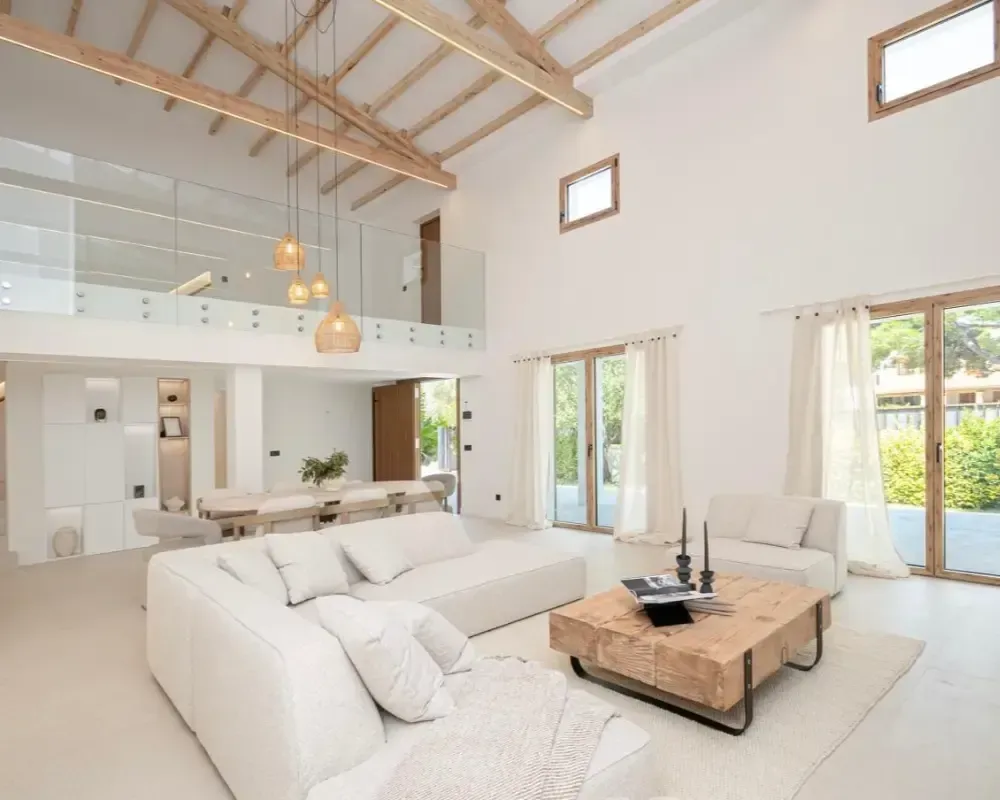 Luminoso salón moderno con sofás blancos, mesa de centro de madera, techo alto y grandes ventanales.