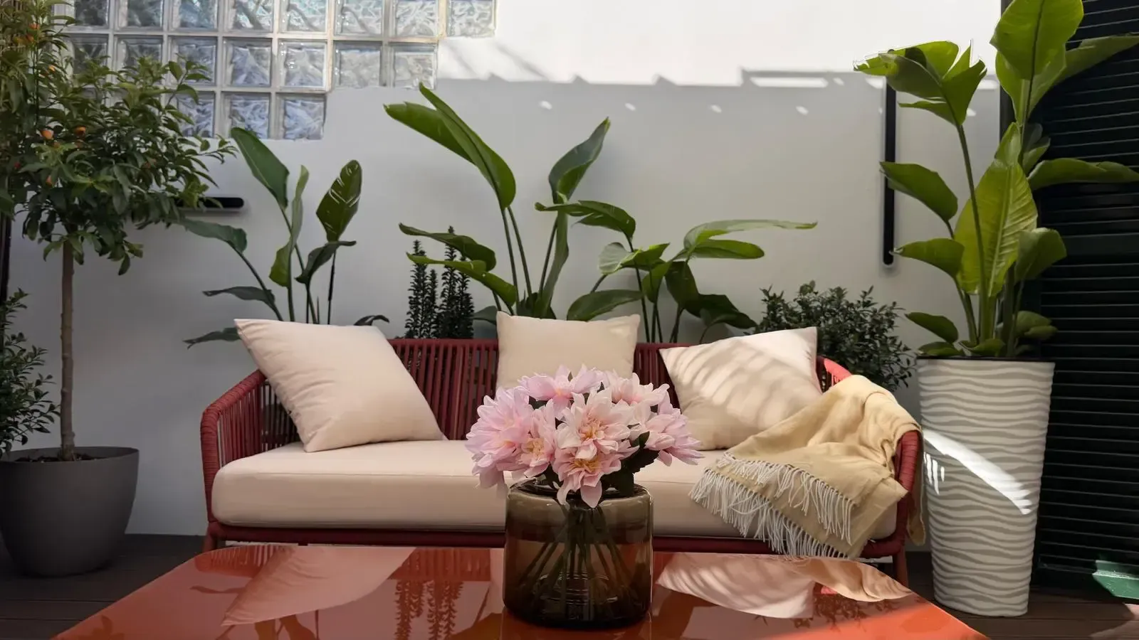 Patio exterior con un sofá rojo, cojines beige, plantas en macetas y una mesa baja con flores rosas.
