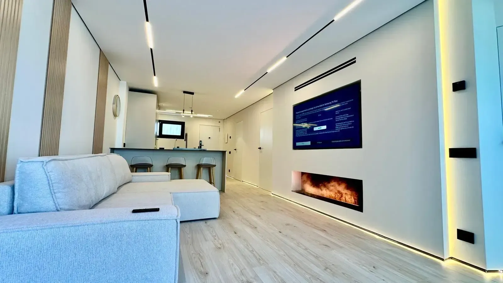 Salón moderno de concepto abierto con sofá blanco, barra de cocina, televisor de pared y chimenea eléctrica.