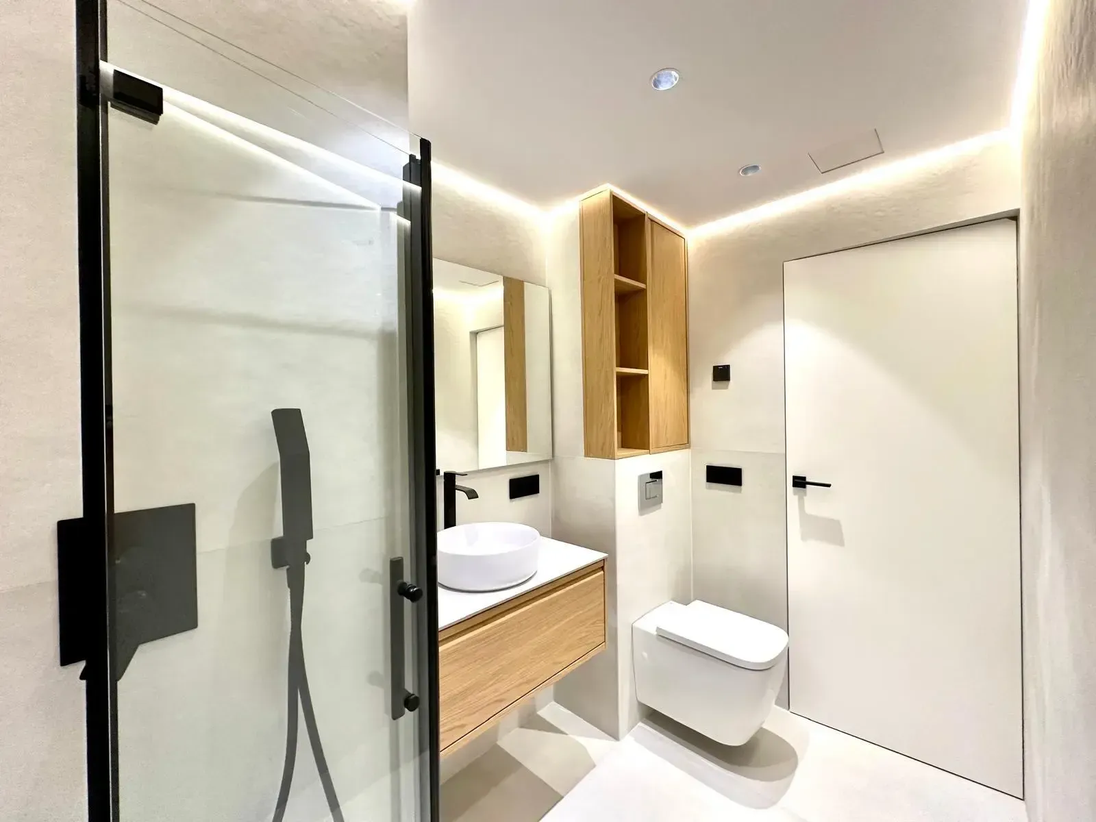 Baño moderno con ducha de cristal, lavabo suspendido, inodoro y estanterías de madera cálida.