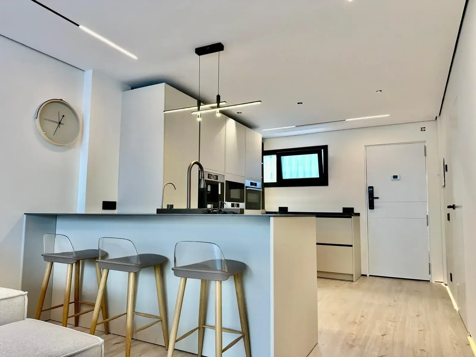 Cocina moderna con armarios blancos, encimeras negras, taburetes de bar y un reloj de pared.