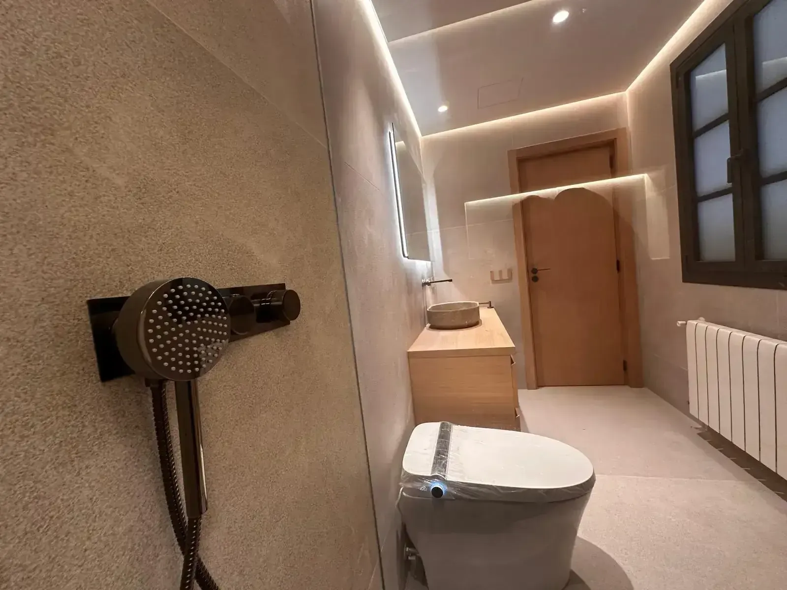 Baño moderno con ducha a ras de suelo, inodoro blanco, lavabo de madera y paredes de azulejos beige.