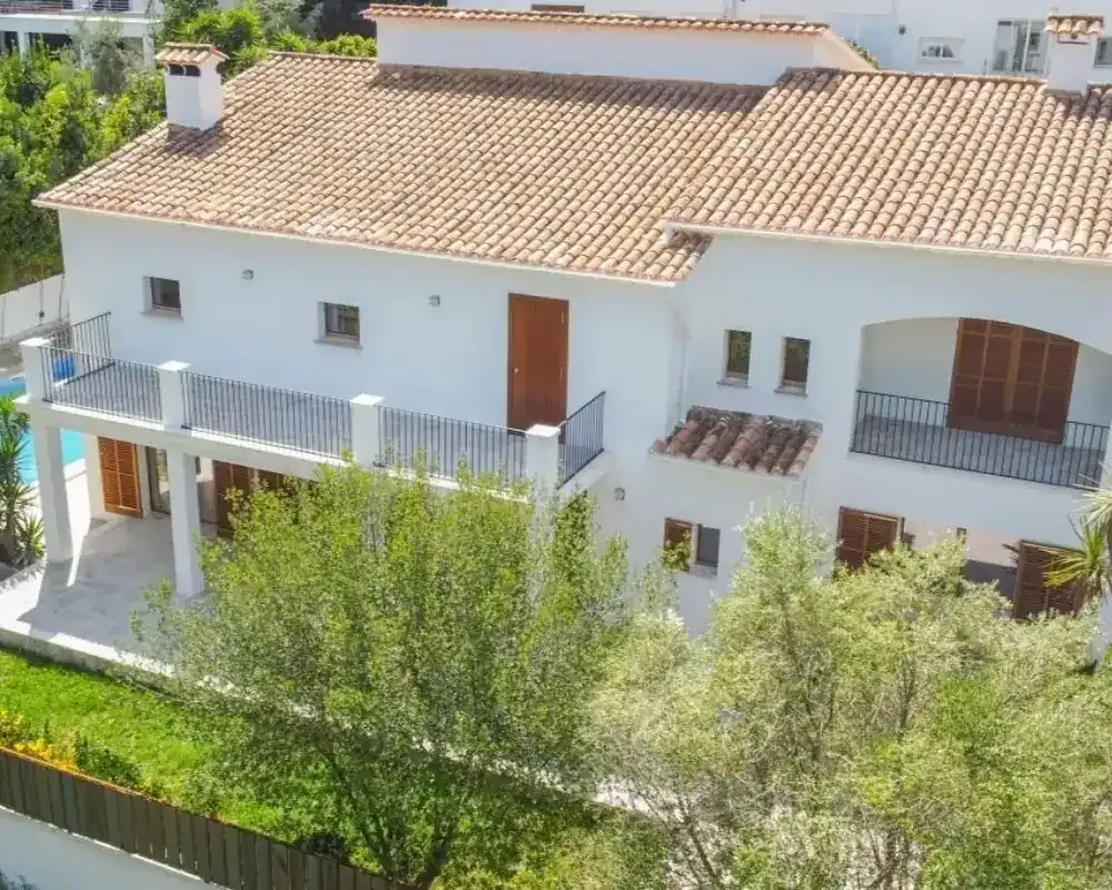 Casa de estuco blanco con techo de terracota, balcones y árboles verdes en la fachada.