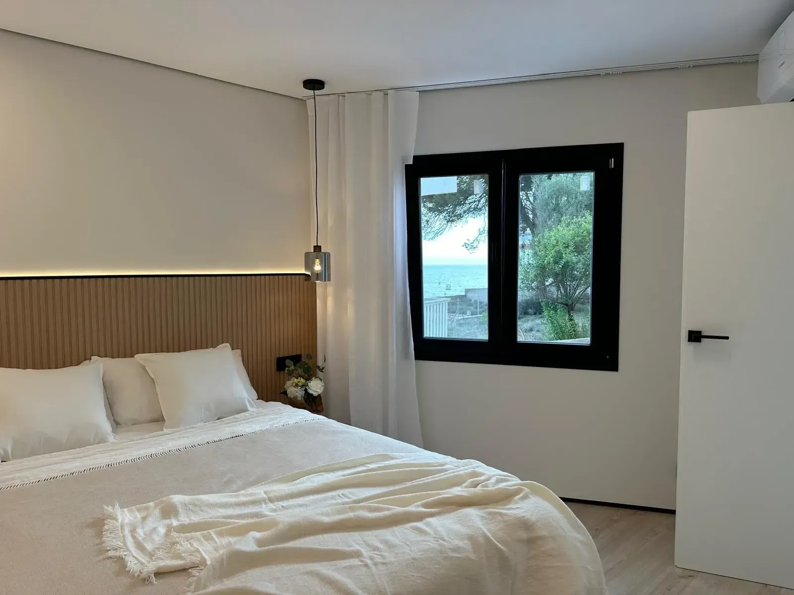 Dormitorio minimalista con ropa de cama blanca, cabecero de madera y una ventana con vistas a árboles y agua.