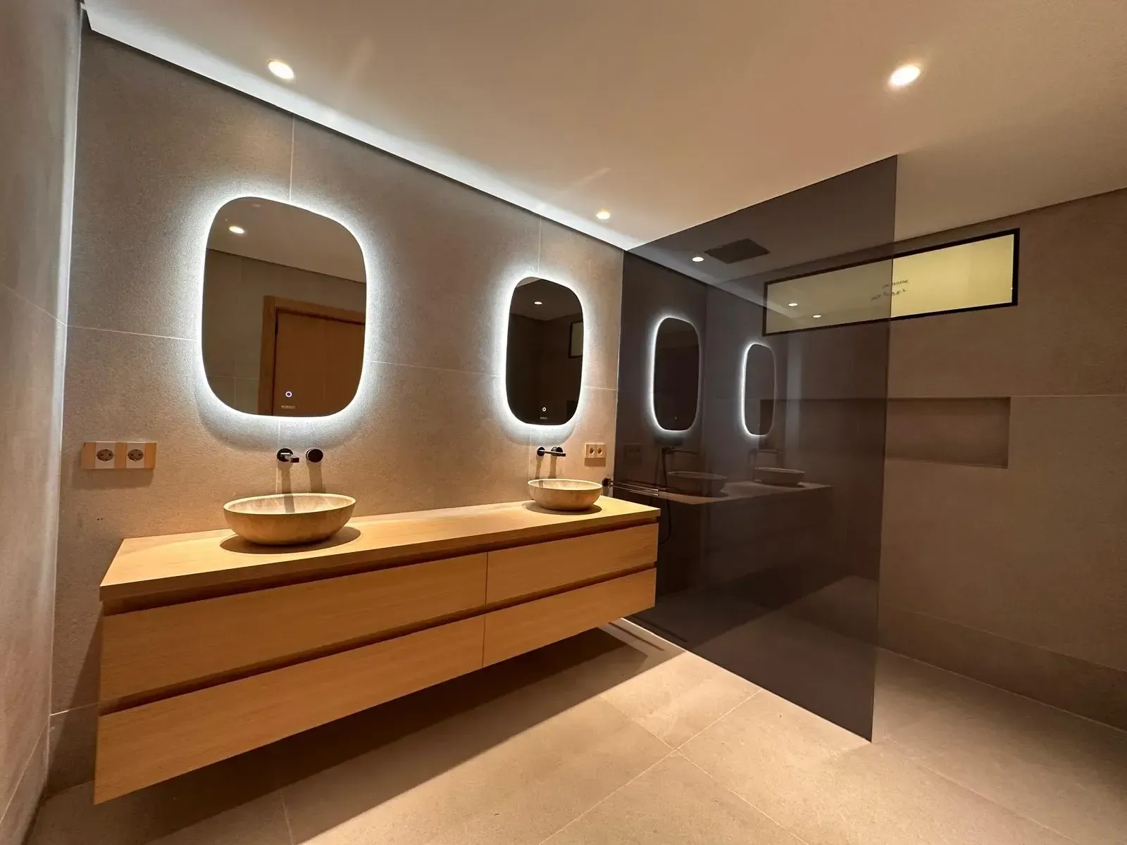 Baño moderno con lavabo doble, espejos retroiluminados y mampara de ducha de cristal.