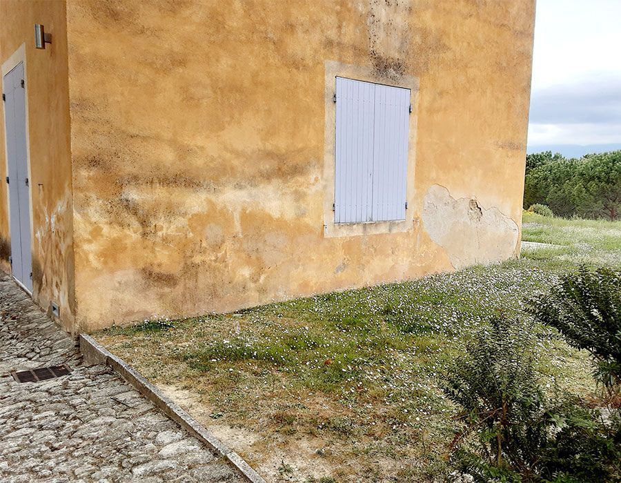 Bâtiment en stuc jaune patiné, aux volets blancs fermés, bordant un chemin de gravier et une cour gazonnée.