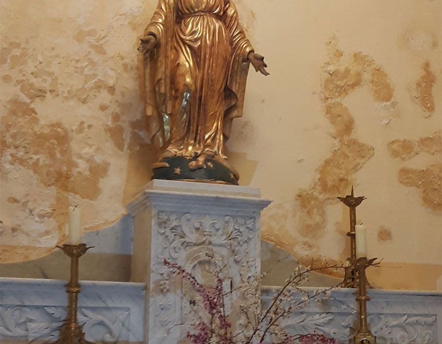 Statue dorée sur un piédestal de marbre dans une niche d'église, flanquée de chandeliers et de peinture murale défraîchie.