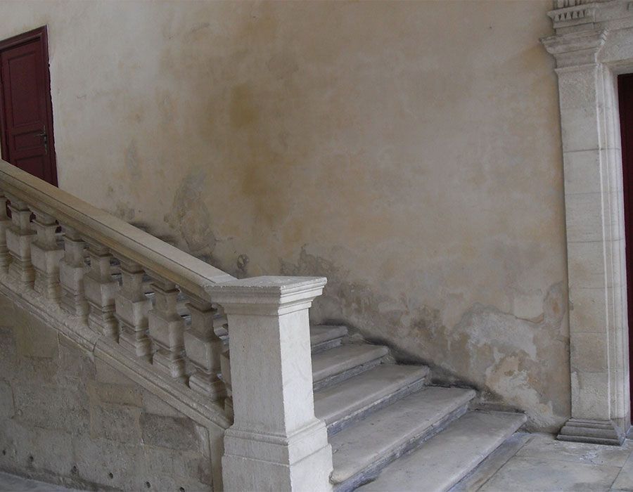 Escalier en pierre poussiéreuse dans un vieux bâtiment avec un mur beige usé et une rampe blanche.