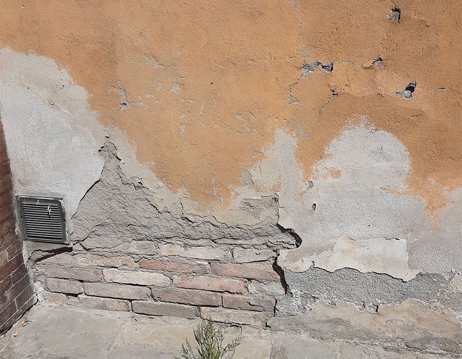 Mur en stuc endommagé avec peinture orange écaillée, briques apparentes et petite grille d'aération près du sol