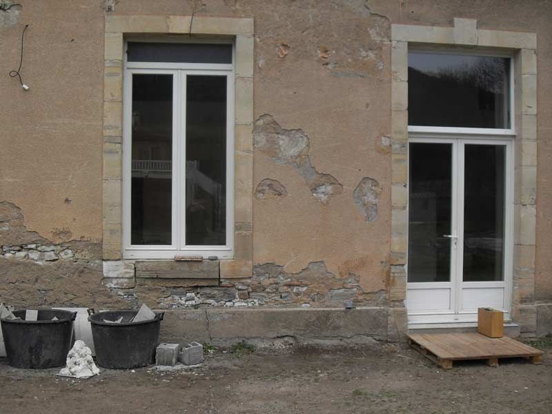 Façade d'immeuble endommagée avec deux fenêtres, plâtre qui s'écaille, poubelles et petite rampe en bois près d'une porte