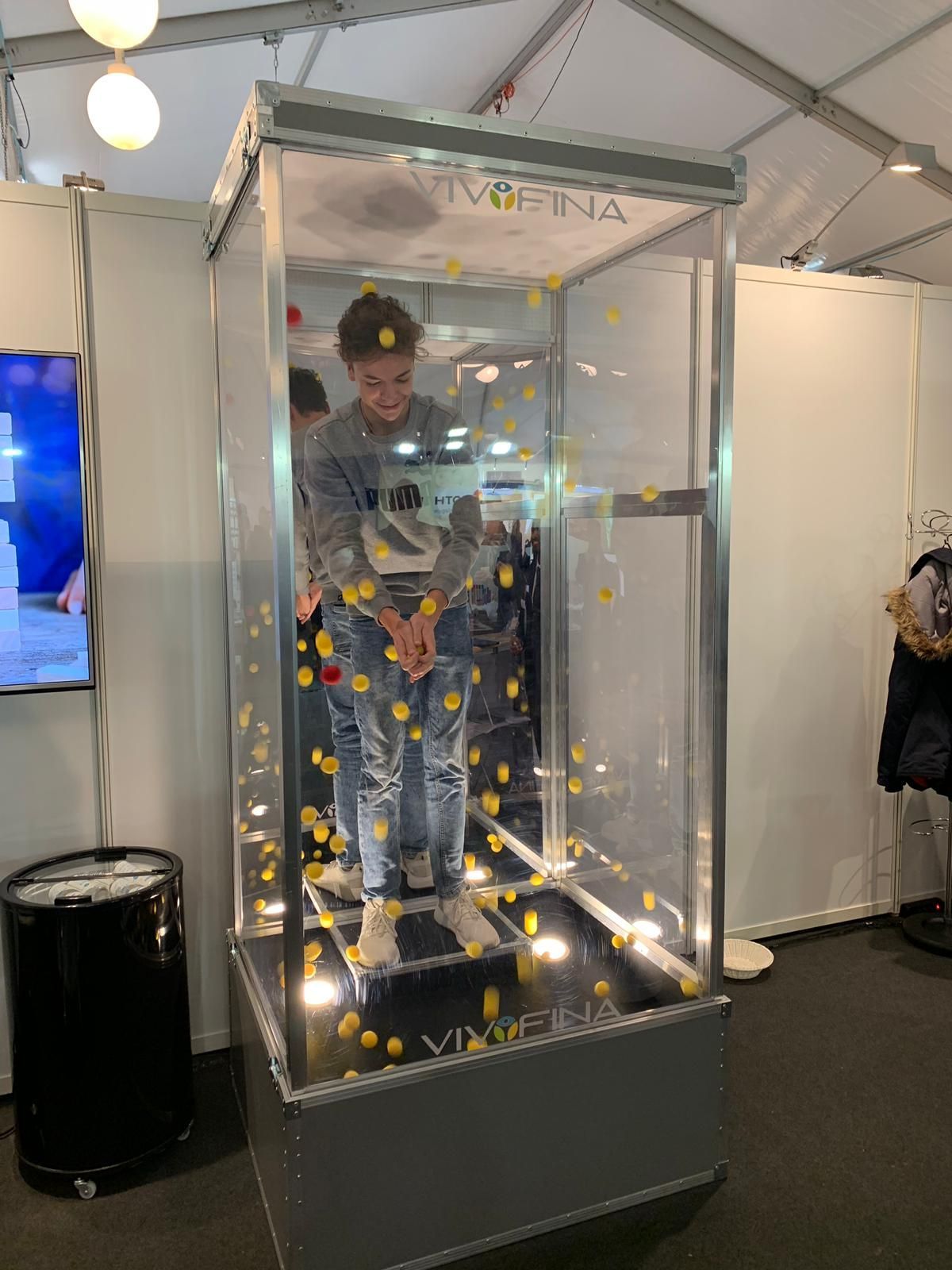 Mann in einer durchsichtigen Kiste mit herabfallenden gelben und roten Kugeln, von unten beleuchtet.