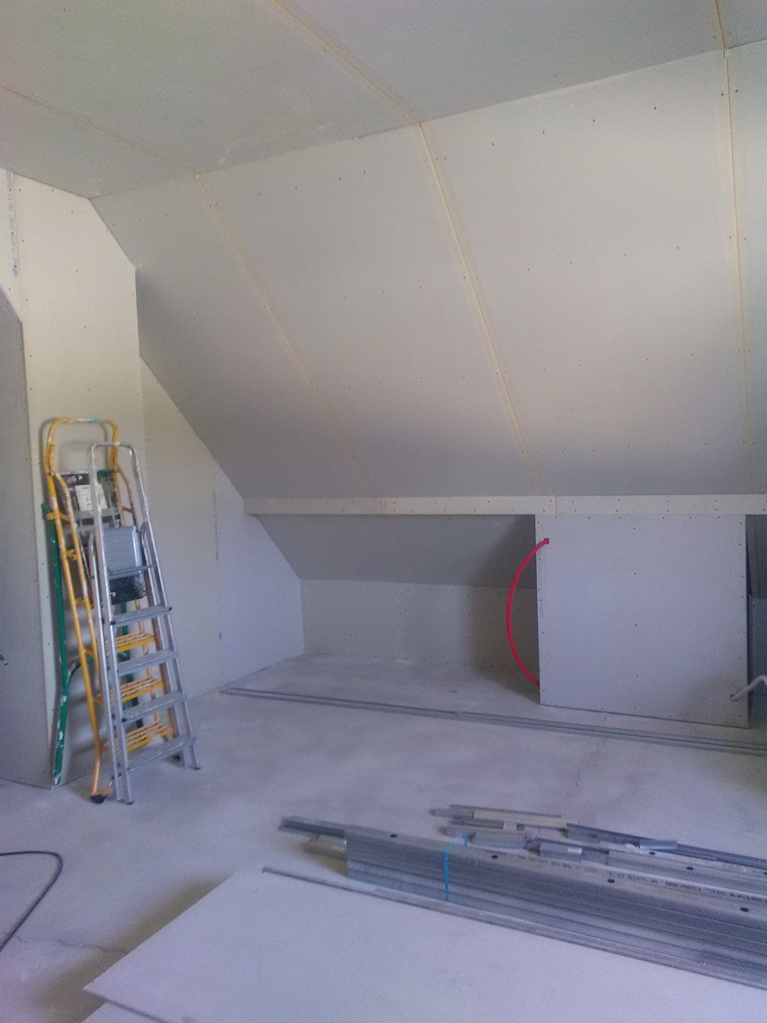 Pièce en rénovation avec plafond mansardé, échelles et poutres métalliques empilées sur un sol poussiéreux.