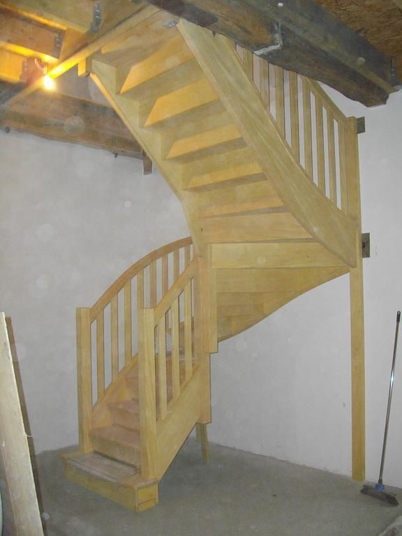 Escalier en colimaçon en bois avec rampe dans une petite pièce intérieure aux murs blancs et aux poutres apparentes.
