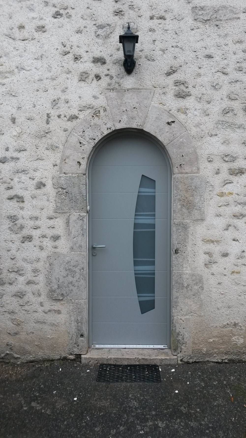 Mur en pierre avec une porte cintrée grise et une petite applique murale noire au-dessus.