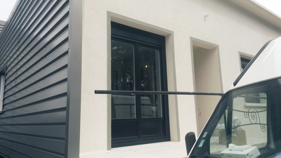 Fenêtre à encadrement noir sur la façade blanche d'un bâtiment, avec un véhicule sombre partiellement visible devant.
