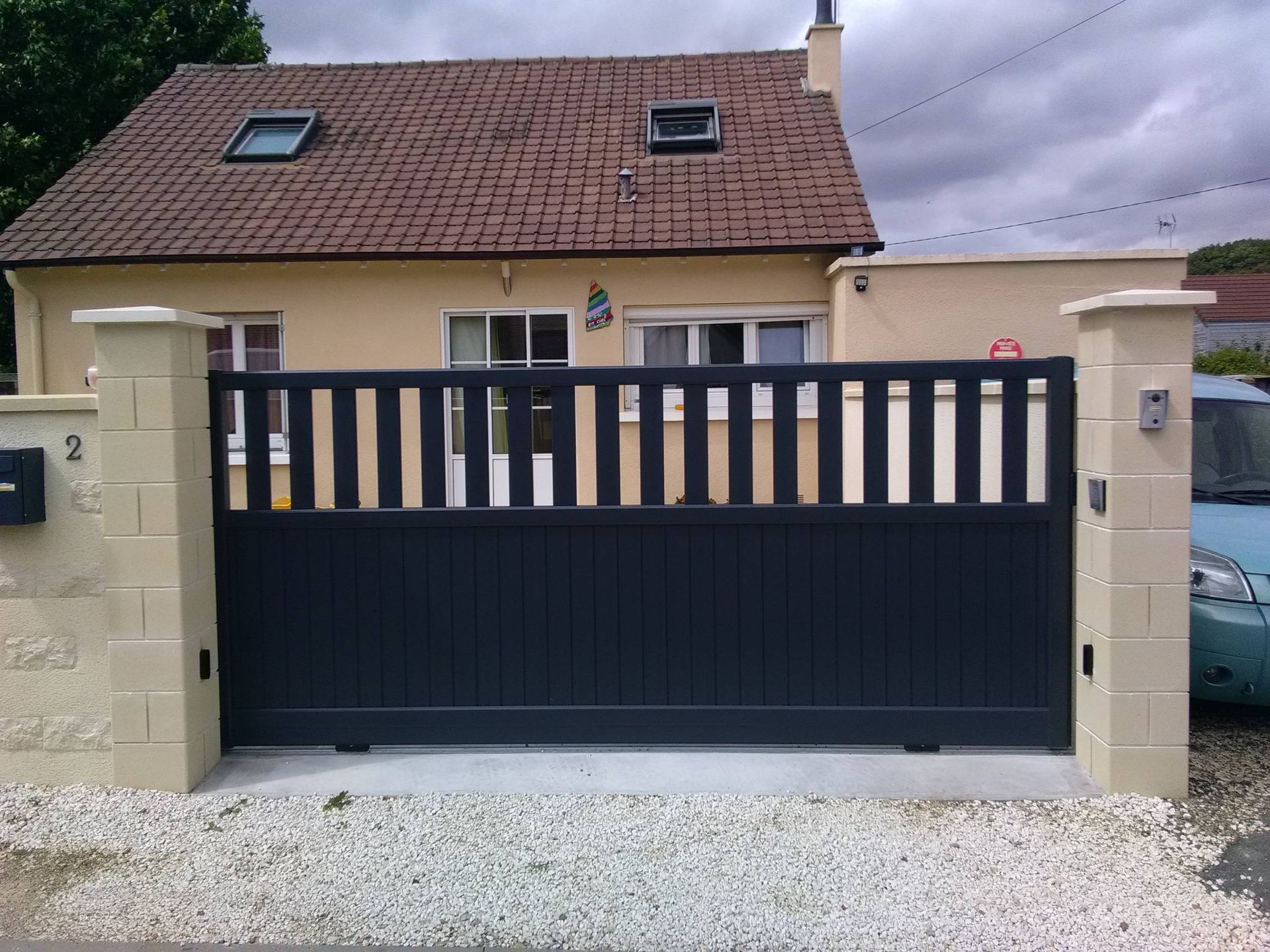 Portail d'allée noir entre des piliers en pierre devant une maison au toit de tuiles et au puits de lumière