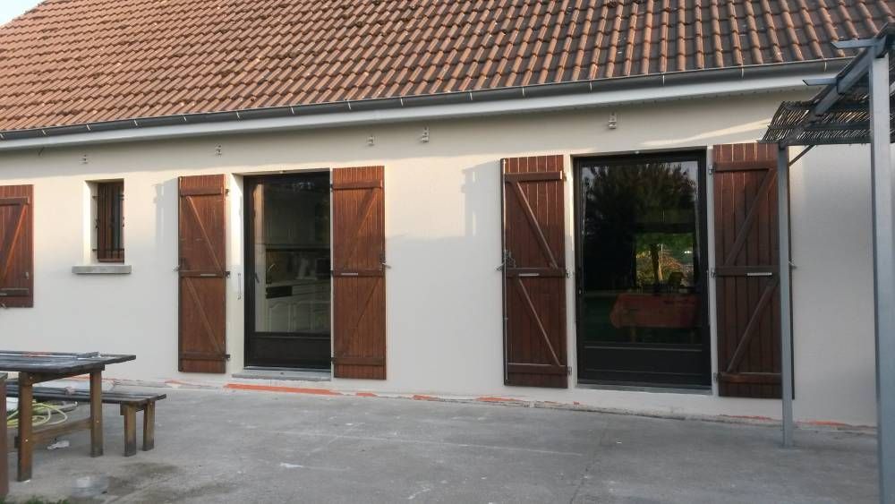 Extérieur d'une maison blanche avec volets bruns ouverts et portes vitrées.