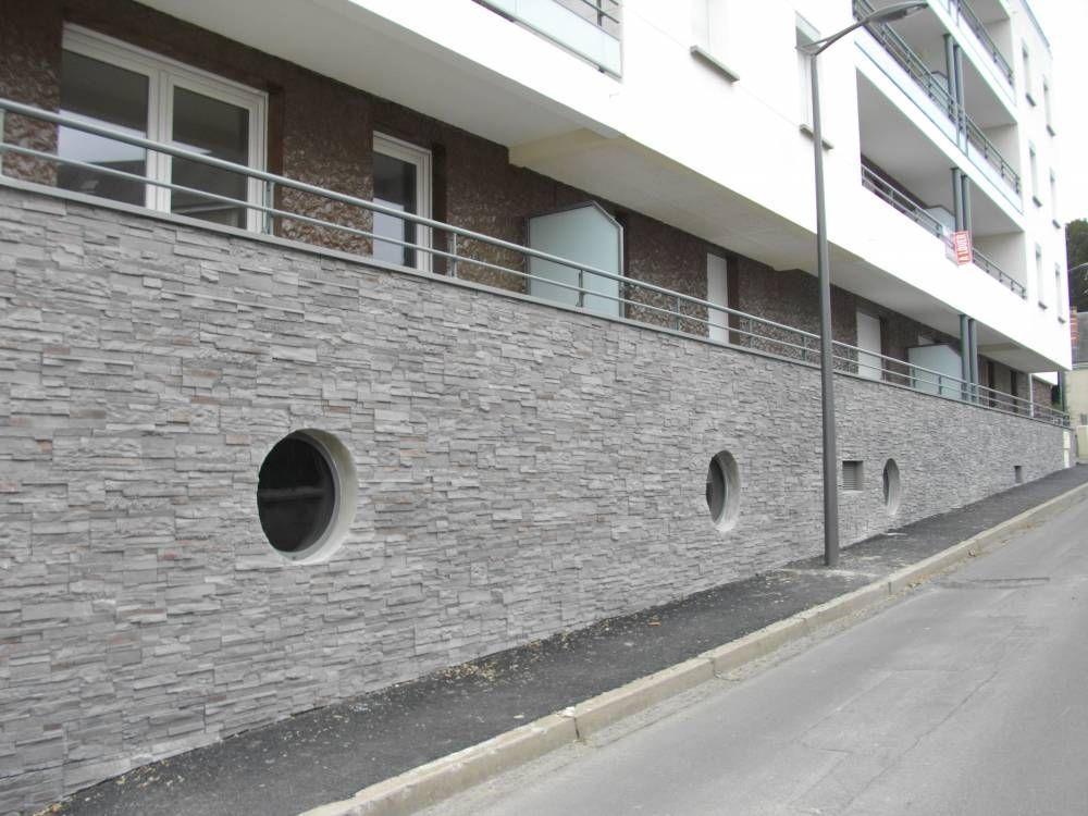 Immeuble d'appartements moderne avec mur en pierre grise et fenêtres circulaires, situé au bord d'une rue en pente.
